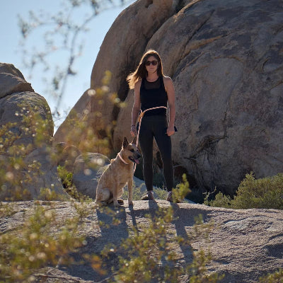 une femme en noir avec des lunettes de soleil debout avec son chien beige attache a sa taille entoures de rochers et de vegetation  nuturelle