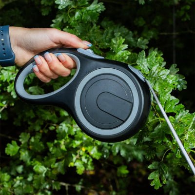 une personne tient une laisse enrouleur grise photographiee en gros plan devant un feuillage vert a l exterieur